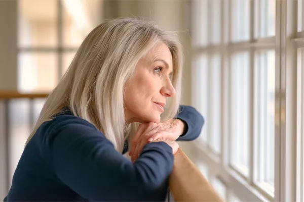 Adrenal-Hormone-Disorder-Clinic A mature woman in a blue shirt looks out the window with melancholy. Schedule treatment for adrenal hormone disorder from Kane Lai, MD in Dallas-Fort Worth Metroplex.