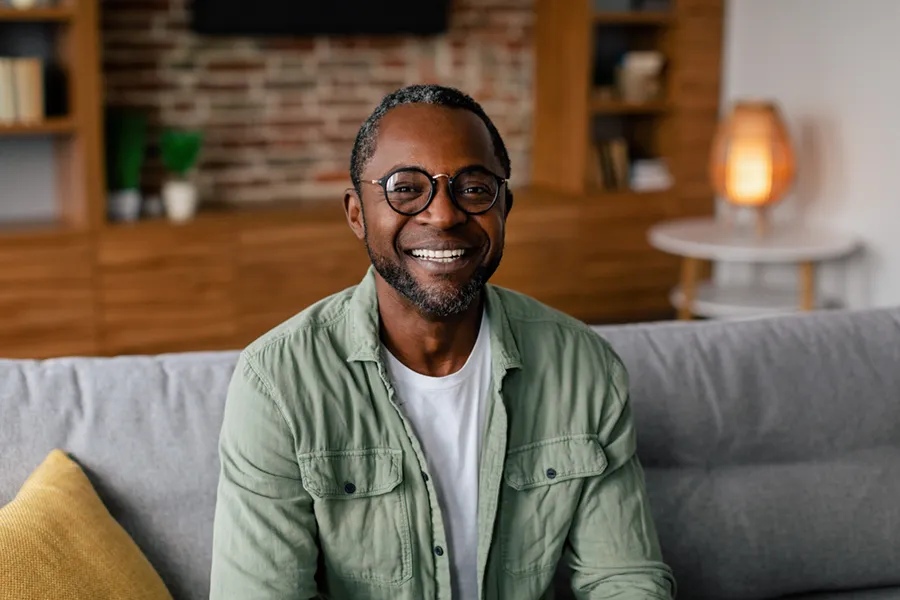 Andropause-Doctor A middle aged black man in a green button up shirt sits on his couch smiling, happy with his treatment for andropause and low testosterone from Kane Lai, MD in Dallas-Fort Worth Metroplex.