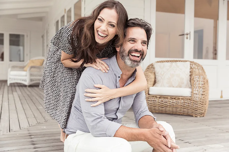 Health-and-Wellness-Services-Doctor A middle-aged couple on a wooden porch with wicker furniture; the woman is playfully hugging the man from behind. They are benefiting from the health and wellness services from Kane Lai, MD in Dallas-Fort Worth Metroplex.