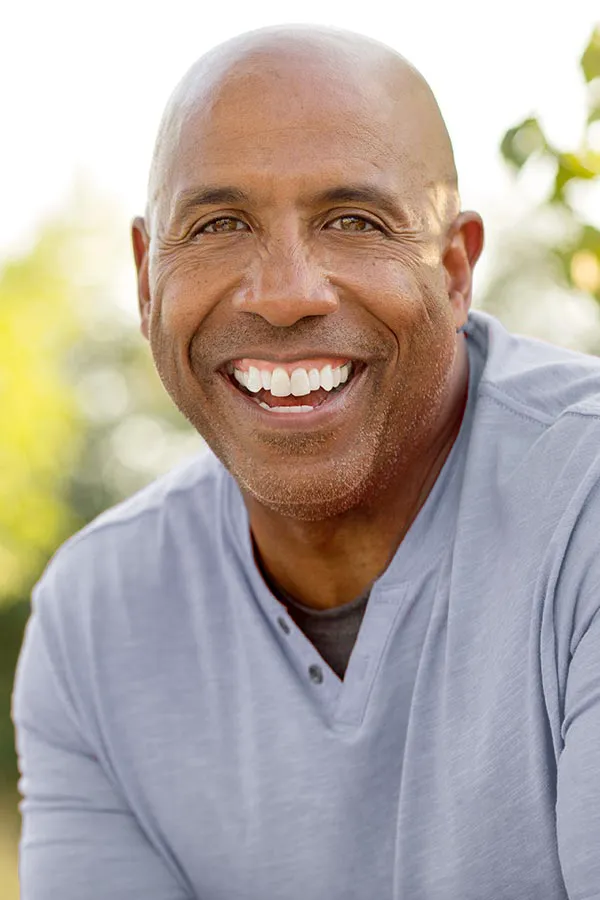 Healthy-Aging-Peptides-Treatment A middle-aged black man in a gray v-neck t-shirt sits outside smiling, happy with the healthy aging peptide treatments from Kane Lai, MD in Dallas-Fort Worth Metroplex