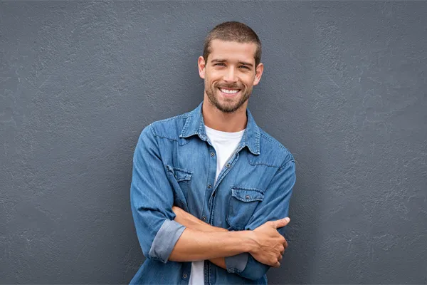 Hormones-for-Men-Doctor A man in a denim shirt stands smiling against a gray-blue wall, pleased with his testosterone hormone therapy from Kane Lai, MD in Dallas-Fort Worth Metroplex.