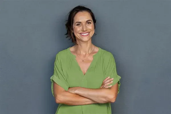 Hormones-for-Women-Doctor A woman in a green shirt stands smiling against a gray-blue wall, pleased with her hormone therapy from Kane Lai, MD in Dallas-Fort Worth Metroplex.