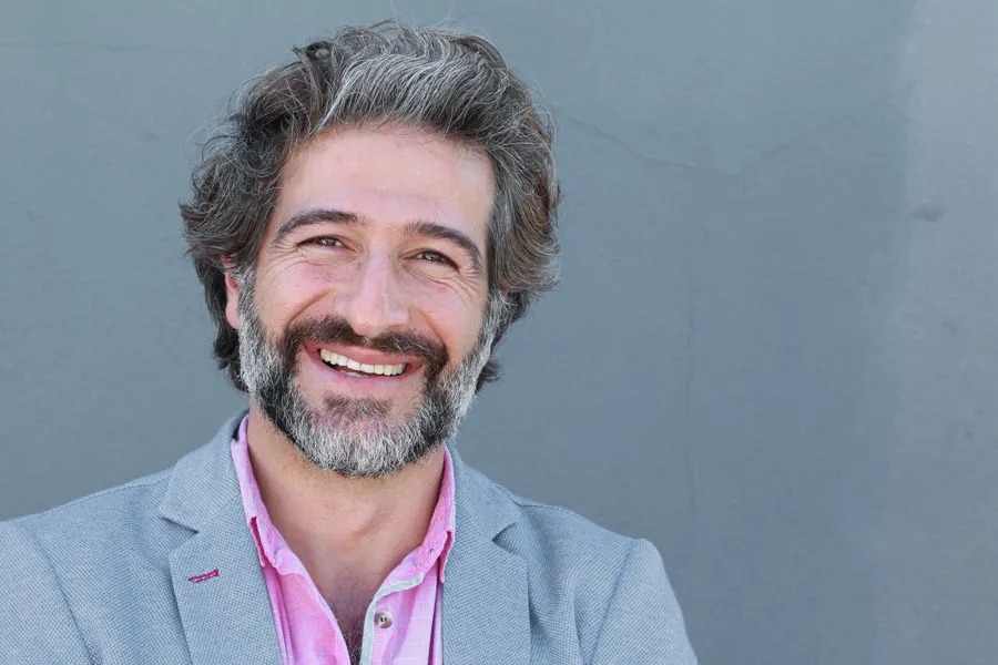 Hypogonadism-Clinic A smiling man with salt and pepper hair and beard wearing a pink shirt and gray blazer stands agains a gray wall. Get hypogonadism treatment from Kane Lai, MD in Dallas-Fort Worth Metroplex.