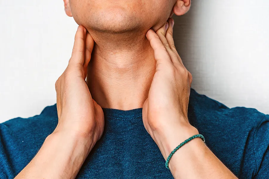 Hypothyroidism-Clinic A man in a dark teal shirt rubs the lymph nodes on his neck. Get hypothyroidism treatment from Kane Lai, MD in Dallas.