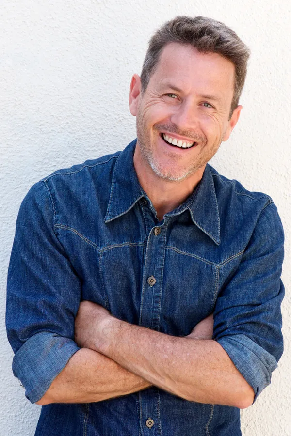 Ipamorelin-Peptide-Treatment A healthy middle-aged man in a denim shirt leans against a white wall smiling. Get Ipamorelin peptide treatment from Kane Lai, MD in Dallas.