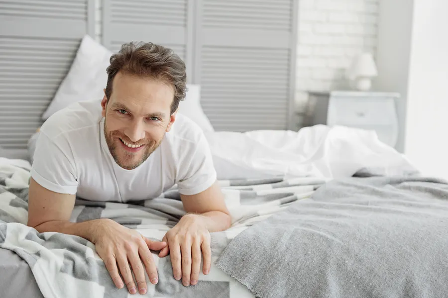 Low-Libido-in-Men-Doctor A dirty-blonde haired man lays on his stomach in bed smiling. Schedule treatment for low libido in men from Kane Lai, MD in Dallas-Fort Worth Metroplex.