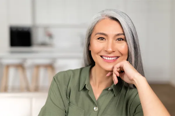 Menopause-Clinic An asian woman with long gray hair and a green shirt sitting and smiling on her couch, happy with her menopause care from [NAME} in Dallas-Fort Worth Metroplex.
