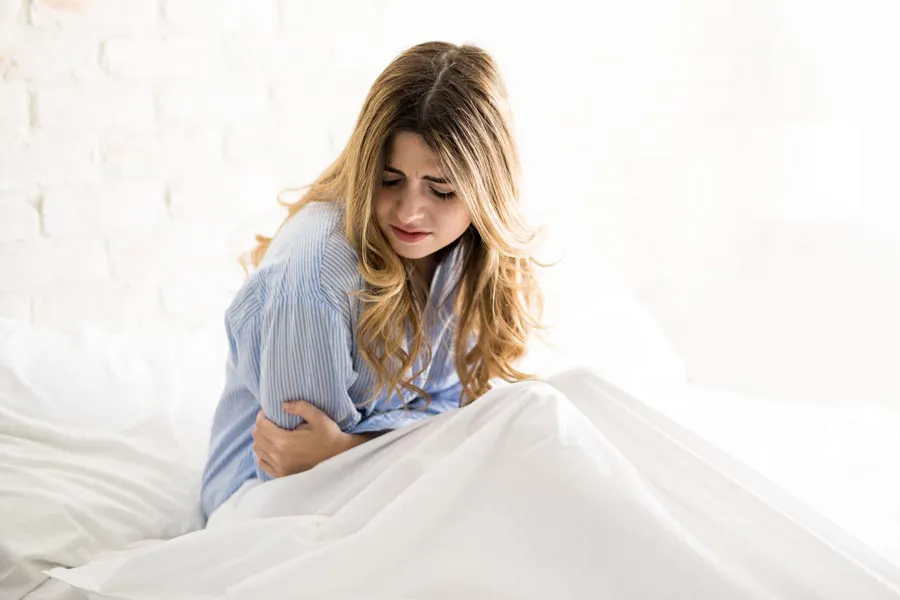 PMS-Therapy A woman with long blonde hair sits in her bed holding her stomach in pain, considering getting care for PMS from Kane Lai, MD in Dallas-Fort Worth Metroplex.