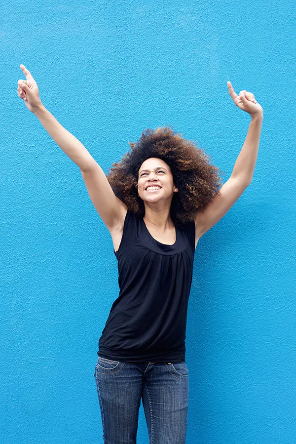 PMS-Treatment A woman in a dark blue tank top standing in front of a bright blue wall, raising her arms in celebration of relief from PMS from Kane Lai, MD in Dallas-Fort Worth Metroplex.