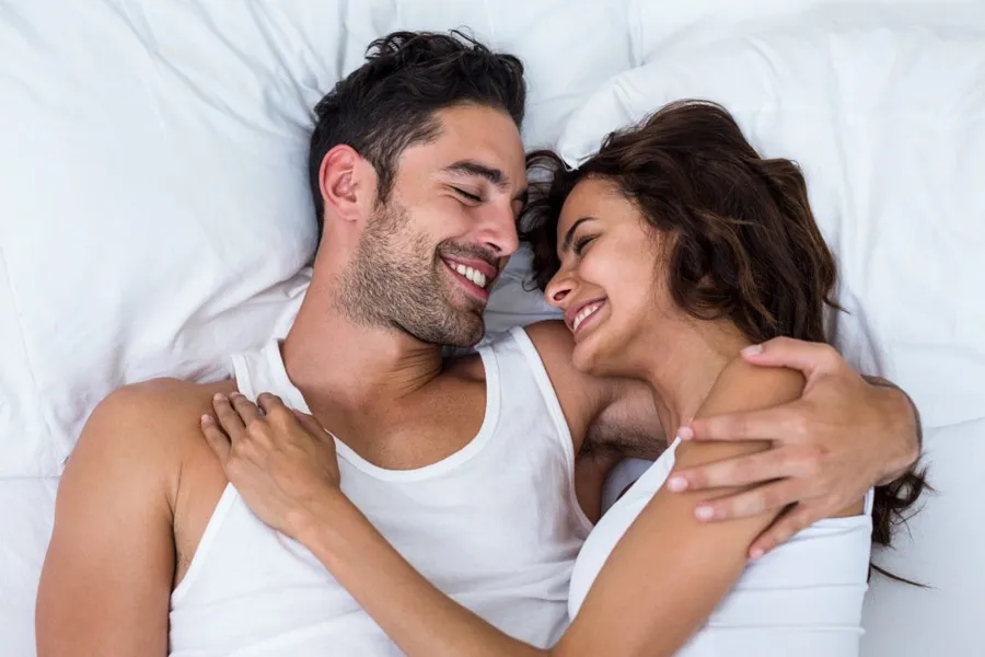 PT141-for-Men-Doctor A laughing man and woman in undershirts lie in each other's arms on a white bed, representing successful PT141 sexual wellness therapy for men from Kane Lai, MD in Dallas-Fort Worth Metroplex.