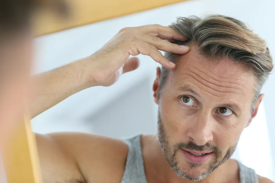Peptides-for-Hair-Restoration-Doctor A middle-aged man with blonde and gray hair inspects his receding hairline in the mirror, considering getting peptides for hair restoration treatments from Kane Lai, MD in Dallas-Fort Worth Metroplex.
