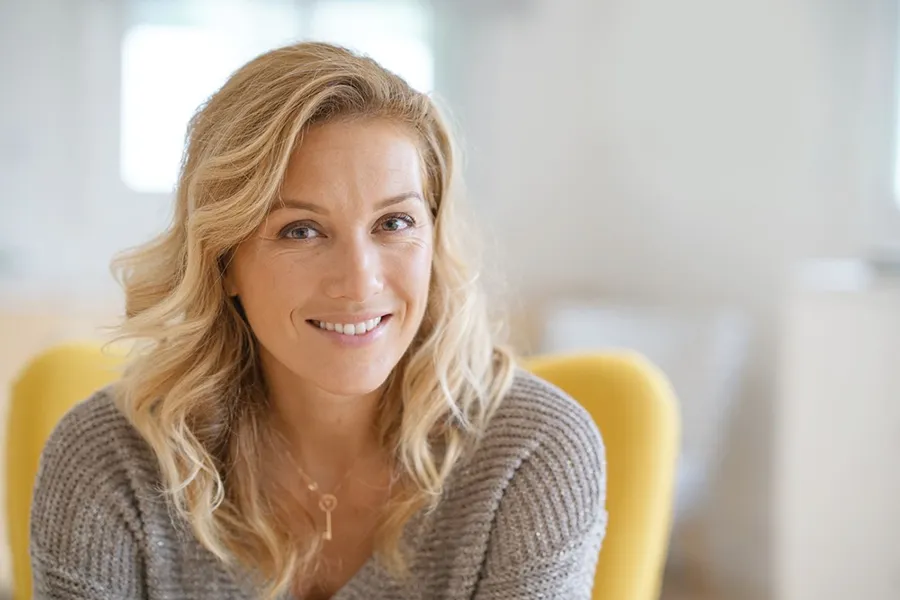 Perimenopause-Doctor A blonde woman wearing a a gray sweater sits inside in a yellow chair, smiling and happy with her perimenopause treatment from Kane Lai, MD in Dallas-Fort Worth Metroplex.