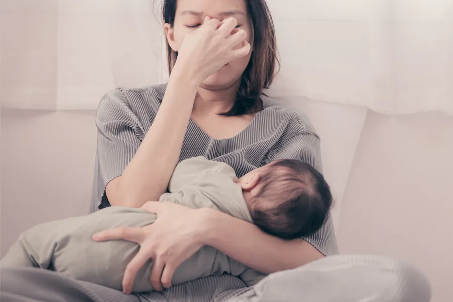 Postpartum-Hormone-Imbalance-Clinic A woman nursing an infant on couch rubs her eyes with exhaustion. Get treatment for postpartum hormone imbalance from Kane Lai, MD in Dallas-Fort Worth Metroplex.