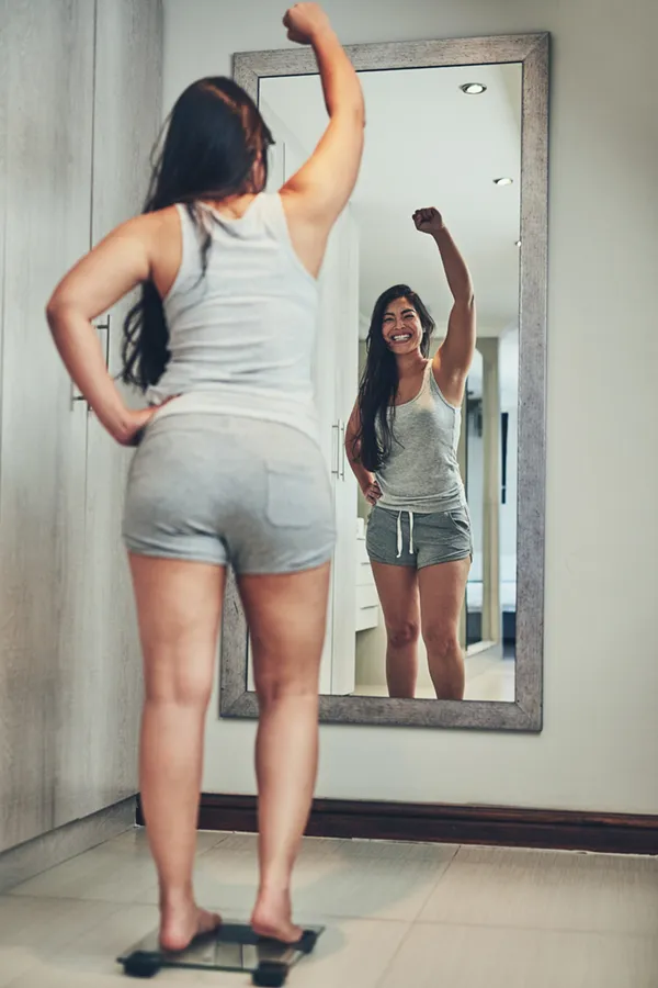 Tirzepatide-Treatment A woman in workout clothes stands on a scale looking in the mirror and pumping her hands in celebration of her successful Tirzepatide treatment from Kane Lai, MD in Dallas-Fort Worth Metroplex.