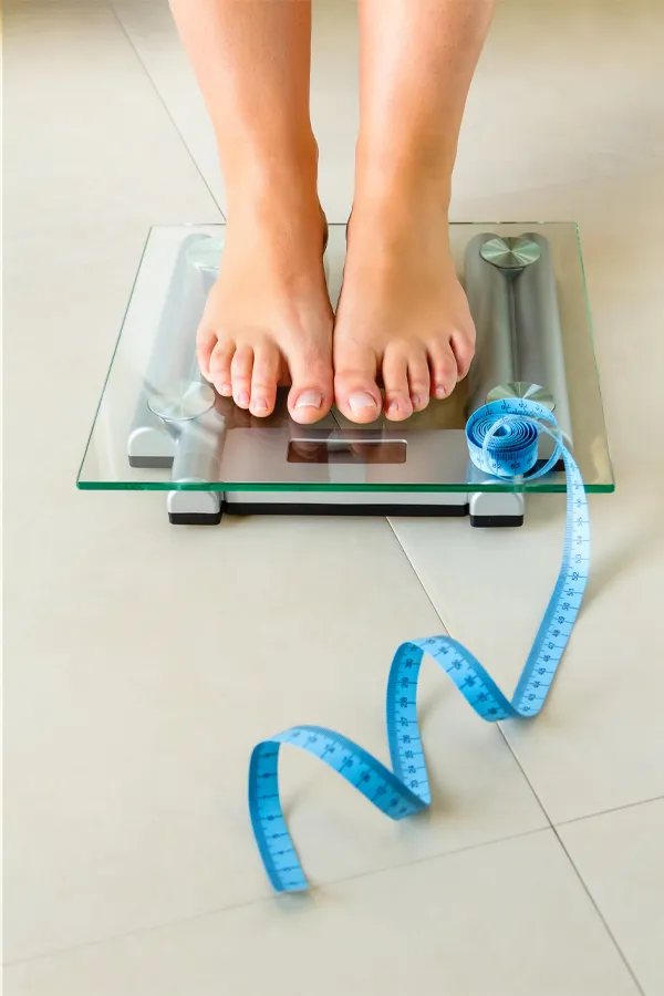 Weight-Loss-Resistance-Treatment Close-up of a woman's feet standing on a scale, with measuring tape by her toes, getting treatment for weight loss resistance from Kane Lai, MD in Dallas-Fort Worth Metroplex.